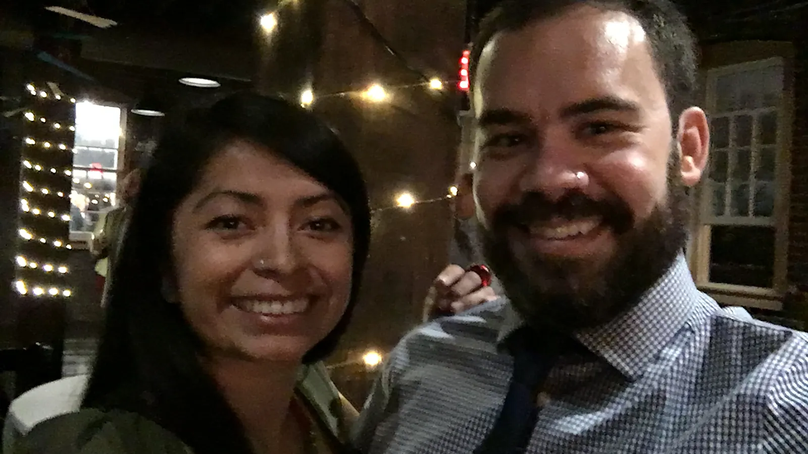 Peter and Consuelo smiling together in a bar hung with string lights