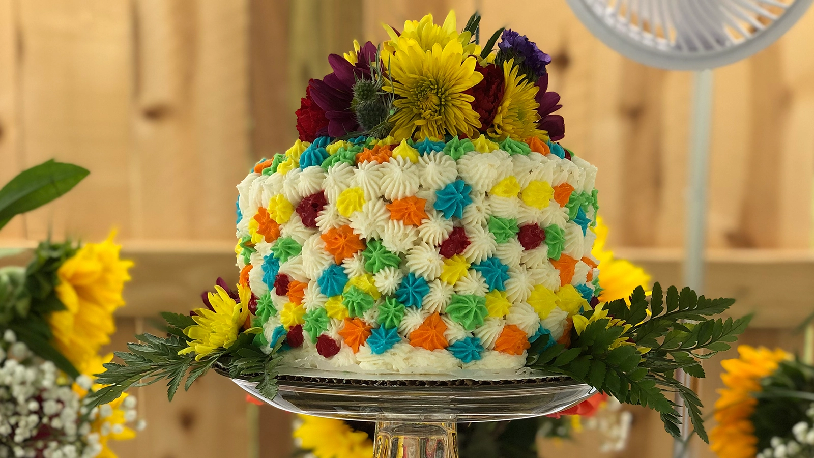 A white cake covered in coloured icing rosettes and topped with fresh flowers