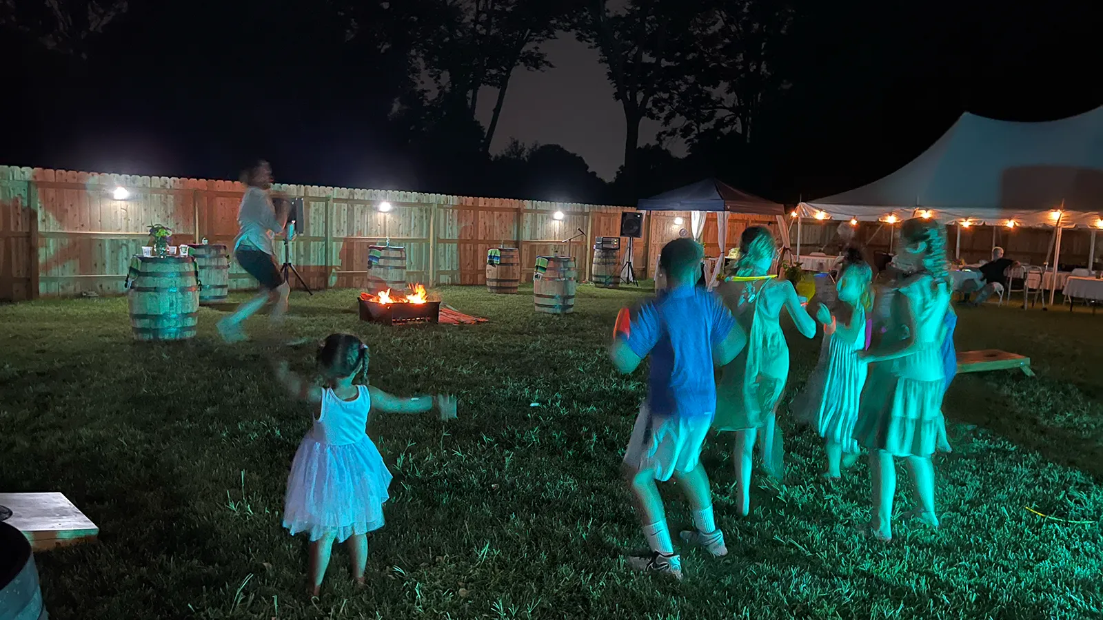 Children dancing on the grass at night, between a fire pit and the lit marquee