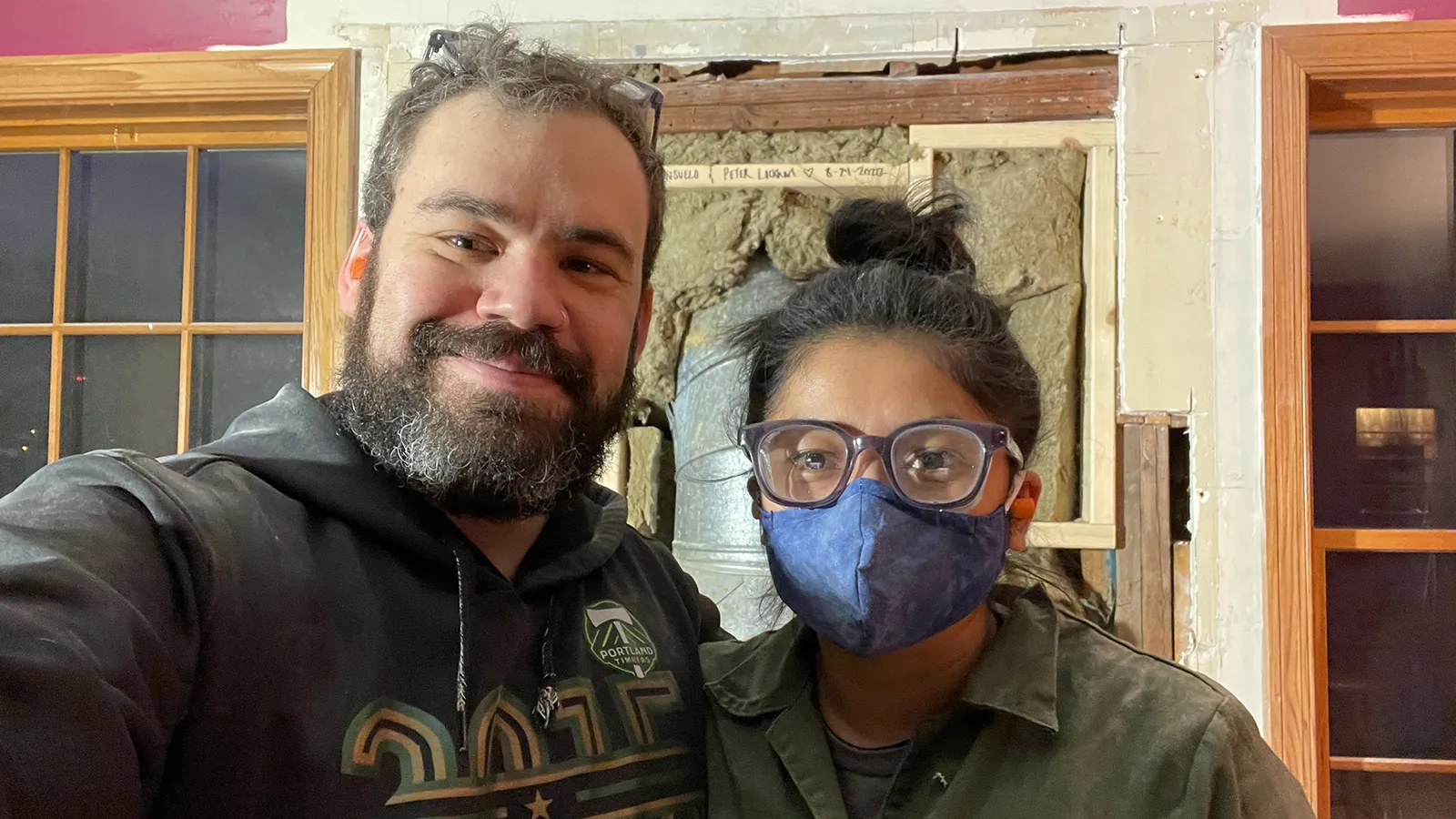 Peter and Consuelo in work clothes in front of an opened-up wall mid-renovation