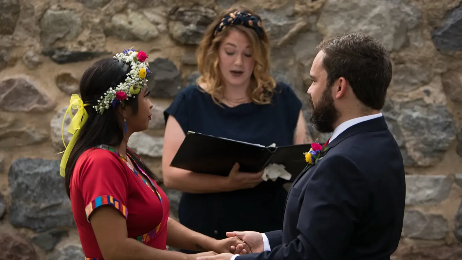 Peter and Consuelo holding hands before the officiant against a stone wall